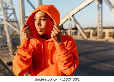 Photo of african american pleased woman in hoodie using earpods while working out on old bridge