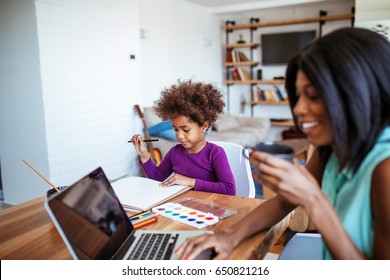 Photo of african american woman working on a laptop while the daughter is painting at the table.