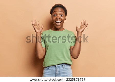 Photo of African woman showcases her elation by raising her palms and exclaiming loudly gets excellent news wears green t shirt and jeans isolated over brown background. Human reactions concept