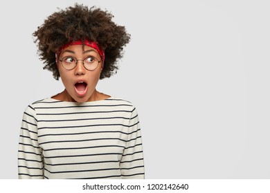 Photo of amazed stupefied black young woman opens mouth widely, looks with stupefaction aside, wears casual clothes, isolated over white background, blank space for your promotional content.