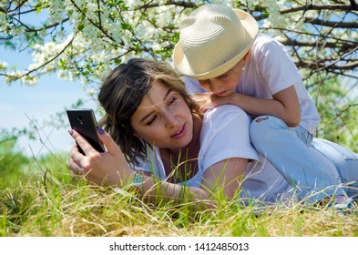 Photo Of An Amazing Young Woman With Her Little Daughter Close-up Outdoors In A Flowered Park, Lying On The Grass, Talking And Looking At A Mobile Phone. Horizontal.