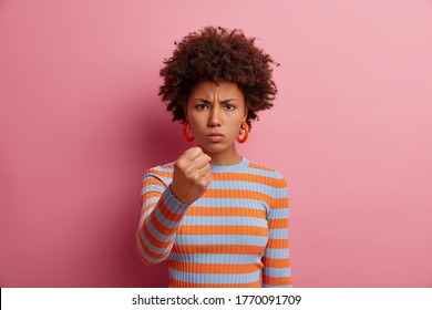Photo of angry hatred woman clenches fist with irritation, expresses aggressive emotions, hate and rage, promises to revenge, wears casual outfit, stands against pink background, has strict face