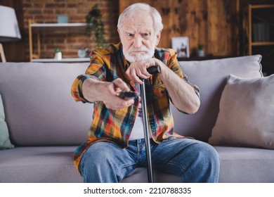 Photo of angry irritated man pensioner wear checkered shirt changing tv channels holding walking cane sitting sofa indoors house room - Powered by Shutterstock