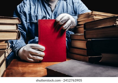 Photo of antiquarian man in a blue shirt and white protective gloves holding a red book and sitting by a table with books pile.