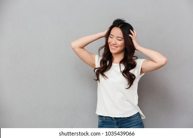 Photo Of Asian Joyous Woman In Casual T-shirt And Jeans Touching Her Beautiful Brown Hair With Candid Smile Isolated Over Gray Background