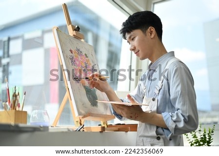 Photo of asian male artist holding paintbrushes and palette painting on easel at bright art studio