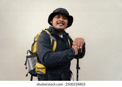 Photo of Asian man wearing adventurer outfit. Half body portrait of an adventurer doing many poses while wearing jacket, hat, backpack and trekking poles isolated on beige background ​