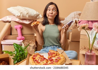 Photo Of Asian Woman Makes Funny Griace Holds Smartphone And Slice Of Pizza Relocates To New Apartment For Living Surrounded By Cardboard Boxes Full Of Personal Belongings. Ownership Concept