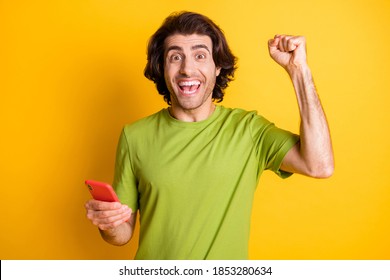 Photo of astonished crazy guy hold telephone raise fist wear green t-shirt isolated yellow color background