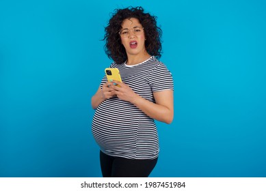 Photo of astonished crazy young Arab pregnant woman wearing stripped T-shirt against blue wall hold smartphone dislike feedback concept - Powered by Shutterstock