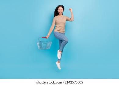 Photo of astonished lady jump hold supermarket basket raise hand wear beige sweater isolated blue color background