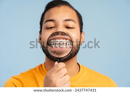 Photo of an attractive African American man with braces, showing his healthy teeth through a magnifying glass, isolated on a blue background. Dental care concept