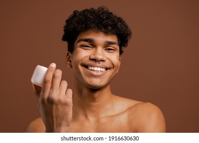 Photo Of Attractive Black Man With Piercing Holds Nutrition Cream And Smiles With White Teeth. Naked Torso, Isolated Brown Color Background