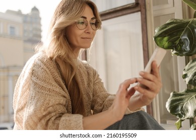 Photo of attractive caucasian adult lady in good mood, holding phone in hands while sitting by window. Blonde woman typing new post in casual warm sweater and glasses. Technology concept