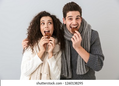 Photo of attractive couple man and woman wearing scarfs eating cookies isolated over gray background