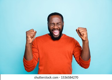 Photo of attractive dark skin guy celebrating lottery winning raising fists excited wear casual orange pullover isolated blue color background
