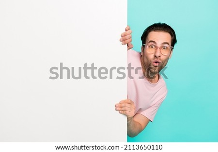 Photo of attractive excited guy wear pink t-shirt hiding behind white wall empty space isolated teal color background