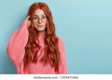 Photo of attractive ginger pupil with long wavy hair, tries to memorize something, keeps fore finger on temple, reminds information for exam, wears spectacles and jumper, stands over blue wall