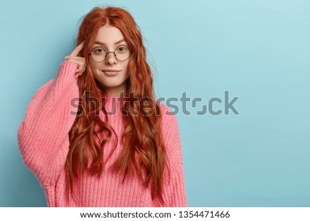 Photo of attractive ginger pupil with long wavy hair, tries to memorize something, keeps fore finger on temple, reminds information for exam, wears spectacles and jumper, stands over blue wall