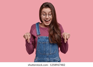 Photo of attractive joyful young European lady with straight dark hair, points down, has happy expression, dressed in casual clothes, models over pink background. Wow, just look at my new shoes!