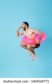 Photo of attractive shirtless tourist man wearing rubber ring smiling while swimming and jumping isolated over blue background