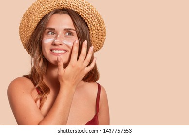 Photo of attractive smiling woman with long hair, has happy facial expression, applaying sunscreen, wearing straw hat, wanting to tan, isolated on beige wall. Summertime, vacation, sunscreen concept.
