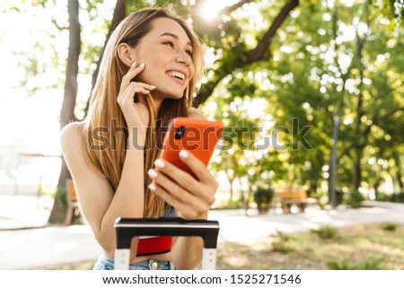 Photo of attractive tourist girl smiling and holding smartphone while sitting with luggage on bench in green park