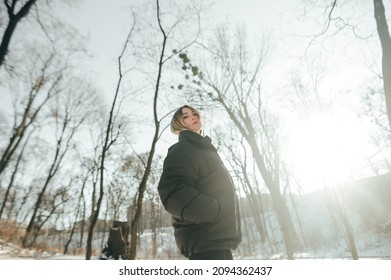 Photo of attractive woman in casual clothes on a walk in the winter forest posing at camera with a serious face on a background of bare trees and the sun
