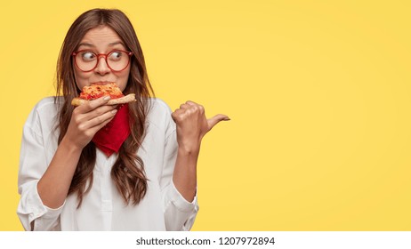 Photo of attractive woman eats slice of pizza, points aside with thumb, dressed in fashionable clothes, shows where pizzeria is, isolated over yellow background. Pretty girl has snack with fastfood