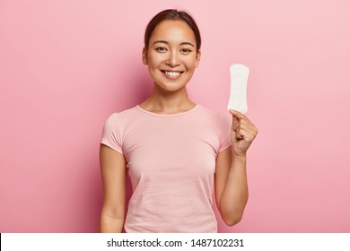 Photo of attractive young lady with Asian appearance, holds clean sanitary napkin, satisfied with its quality, uses intimate product during menstruation or periods, isolated on pink background