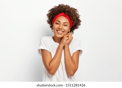 Photo of attractive young woman with Afro hairstyle, keeps hands pressed together, wears red headband, casual clothes, poses against white background, has toothy smile on face, gets compliment