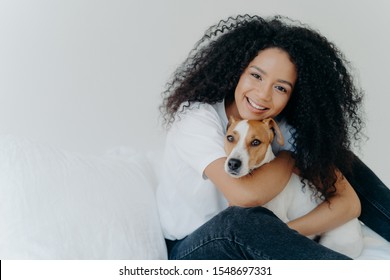 Photo Of Attractive Young Woman With Afro Hircut, Embraces With Love Dog, Takes Care Of Pet, Smiles Gently, Wears Casual Clothing, Isolated Over White Background, Sit On Bed, Copy Space For Your Promo