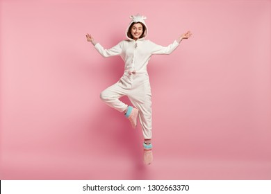 Photo of attractive young woman jumps high dressed in soft kigurumi, meditates indoor, jumps indoor, isolated over pink studio background, tries to relax, awakes in morning. Happiness concept