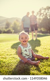 Photo Of The Baby And His Grandparents In The Background. Smiling Cute Mixed Race Baby Boy Sitting On The Grass Outdoors Being Watched By His Grandparents