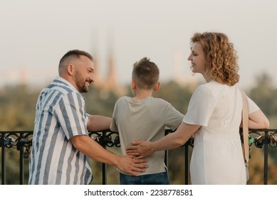 A photo from the back of father, mother, and son who are staring at the old European church. The happy family of tourists is enjoying the town sight in the evening. Tourists at sunset. - Shutterstock ID 2238778581
