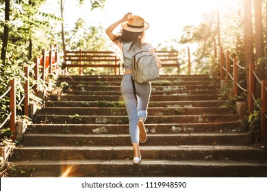 Photo from back of young woman 18-20 wearing summer clothes and straw hat running up stairs in green park on sunny day