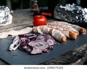 Photo of bacon wrapped sausages hot dogs with tomato onion and a hunting knife outdoor grill picnic food preparation