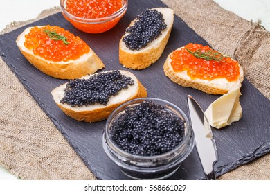 Photo of baguette with black and red caviar, knife, cup on blackboard