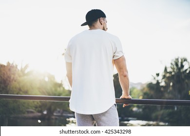 Photo Bearded Muscular Man Wearing White Blank t-shirt, snapback cap and shorts in summer time. Green City Garden Park Sunset Background. Back view. Horizontal Mockup.