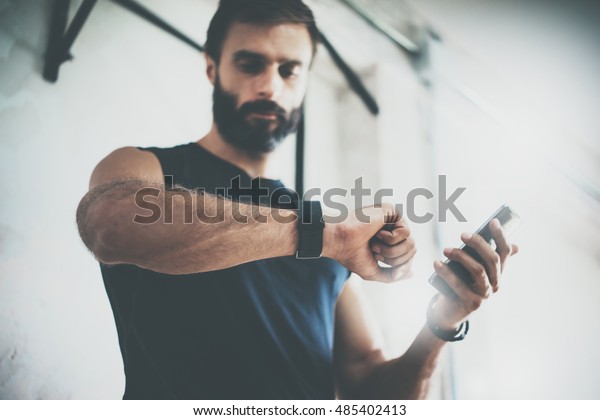 Photo\
Bearded Sportive Man After Workout Session Checks Fitness Results\
Smartphone.Adult Guy Wearing Sport Tracker Wristband Arm.Training\
hard inside gym.Horizontal bar\
background.Blurred