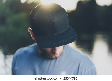 Photo Bearded Young Man Wearing Black Blank Cap. Green City Park Lake Background and Sunset effect. Front view. Horizontal Mockup.