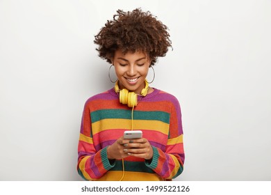 Photo of beautiful Afro American woman concentated into smartphone device, enjoys chatting online, downloads music to playlist for listening in headphones, has curly dark hair, wears casual outfit