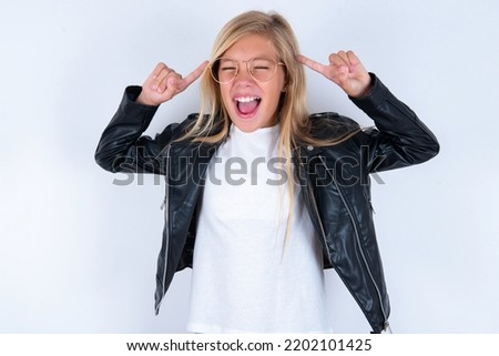 Photo of beautiful caucasian blonde little girl wearing biker jacket and glasses over white background screaming and pointing with fingers at hair closed eyes