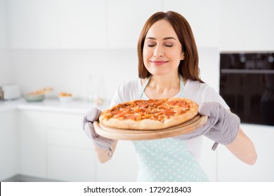Photo of beautiful cheerful housewife enjoy hobby cooking family recipe dinner pizza smelling hot ready meal from oven wear t-shirt apron gloves standing modern kitchen indoors