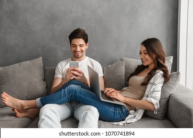 Photo Of Beautiful Couple Man And Woman 30s Resting In Living Room On Couch And Using Laptop Computer And Mobile Phone