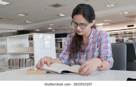 Photo of beautiful and intelligent young girl with eyeglasses studying hard in public library for upcoming exams. Every college going student may connect to this picture for various scenarios.