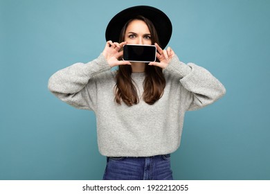 Photo of Beautiful positive young female person wearing black hat and grey sweater holding mobilephone showing smartphone isolated on background looking at camera