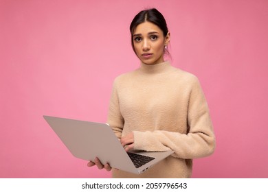 Photo Of Beautiful Sad Gloomy Young Brunette Woman Wearing Beige Sweater Holding Netbook Computer Typing Text On Keyboard Looking At Camera Isolated Over Pink Wall Background