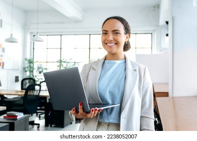 Photo of beautiful successful brazilian or latino business woman, in elegant clothes, seo, top manager, programmer, stands in a modern office, holding an open laptop in hands, looks at camera, smiling