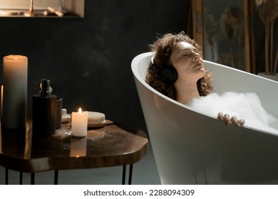 Photo of a beautiful woman who lies in the bath with her eyes closed and listens to music through wireless headphones. A young woman relaxes in a bath with aromatic candles and listens to music on - Powered by Shutterstock